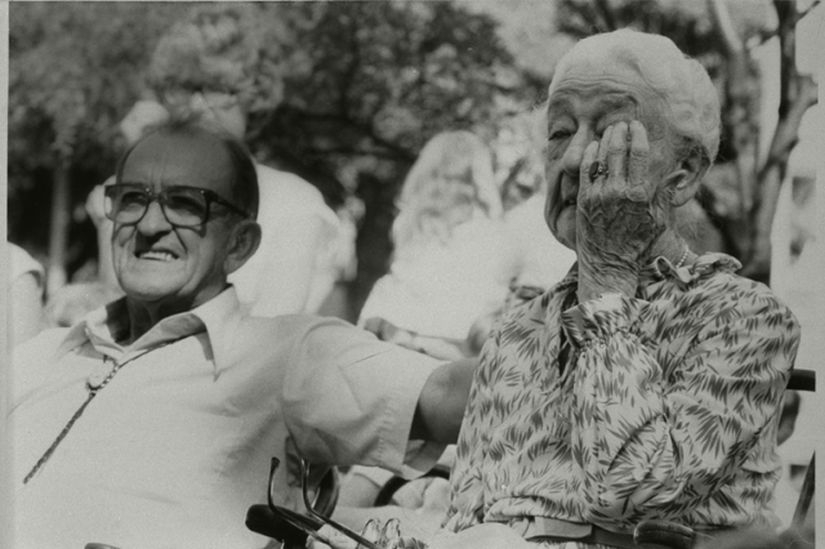 An older woman with her glasses off to wipe her eyes with a smiling man seated next to her.