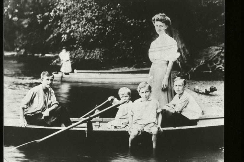 Four boys of varying ages sitting in a row boat with a young women standing behind them in the boat. A young girl in a boat in the background.