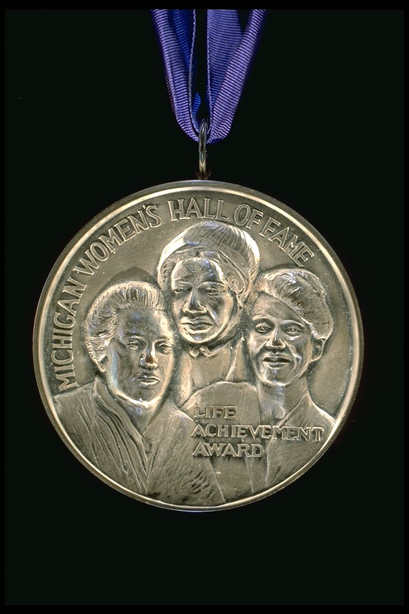 Silver medal on a blue ribbon with three women with the words "Michigan Women's Hall of Fame along the top and the words: Life Achievement Award" near the bottom. 