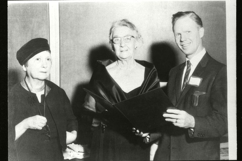 A man holding a folder open for a women to look at with another woman looking on. 